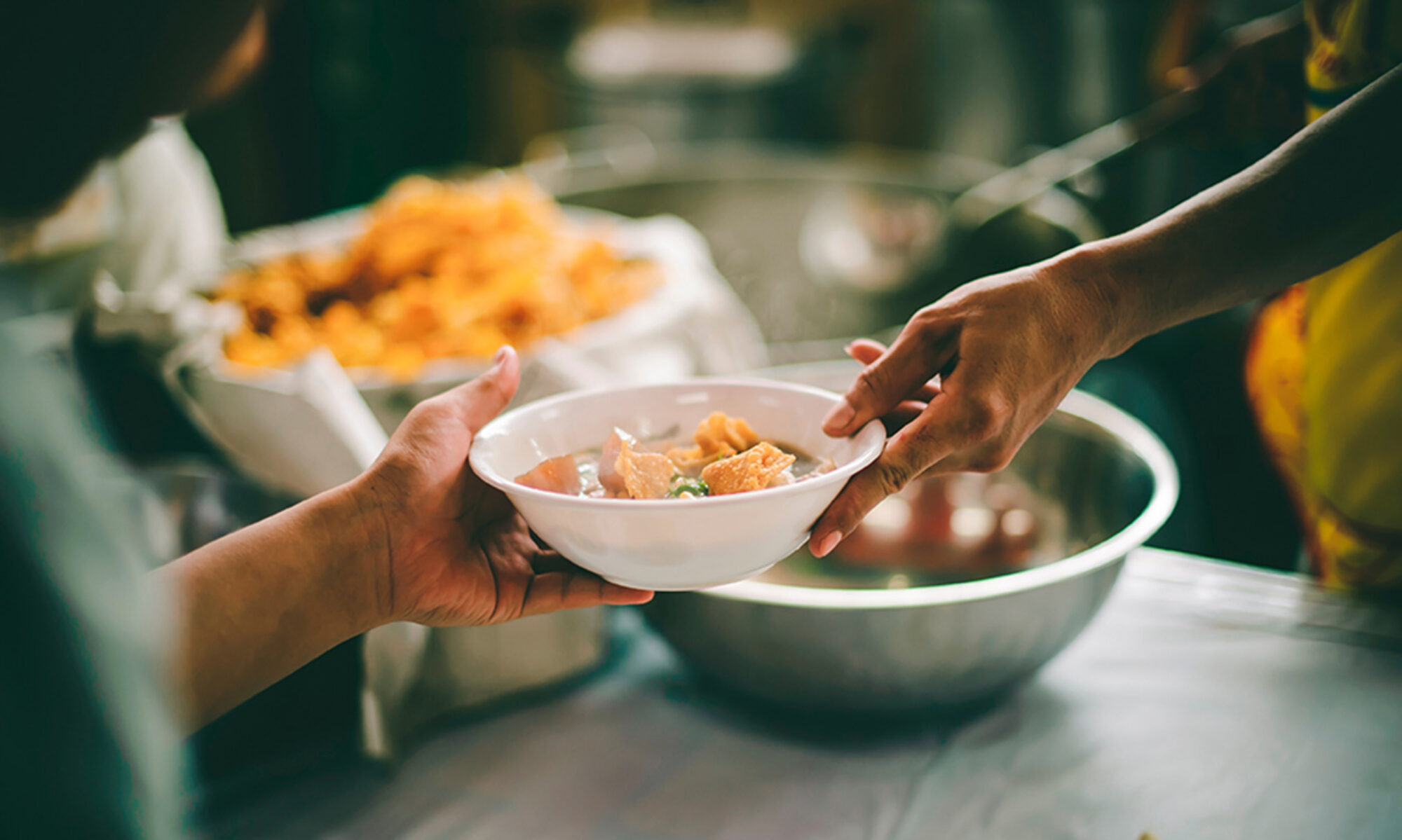 A photo of a person handing a bowl of food to another person. There is food in the background