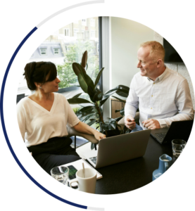 An image of employees in business attire speaking with each other in an office. They are sitting next to each other with their laptops open