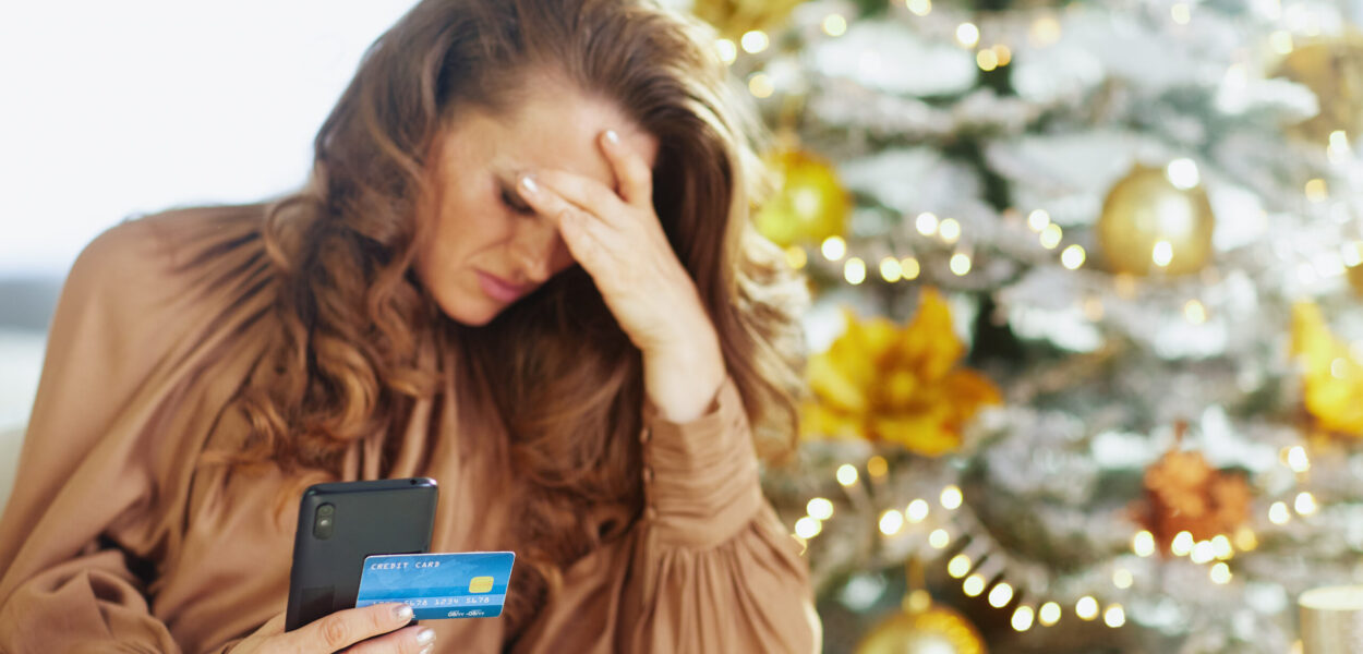 A stressed woman holds smartphone and credit card, reflecting on financial anxiety. The blurred Christmas tree in background highlights concerns about overspending or budgeting during festive season