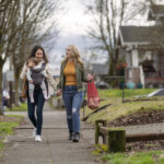 A young mixed race lesbian couple hold hands while walking on a sidewalk through an urban residential neighborhood. One woman is carrying a one year old baby in an ergonomic baby carrier while the other woman carries groceries in sustainable reusable shopping bags.