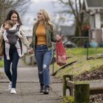 A young mixed race lesbian couple hold hands while walking on a sidewalk through an urban residential neighborhood. One woman is carrying a one year old baby in an ergonomic baby carrier while the other woman carries groceries in sustainable reusable shopping bags.