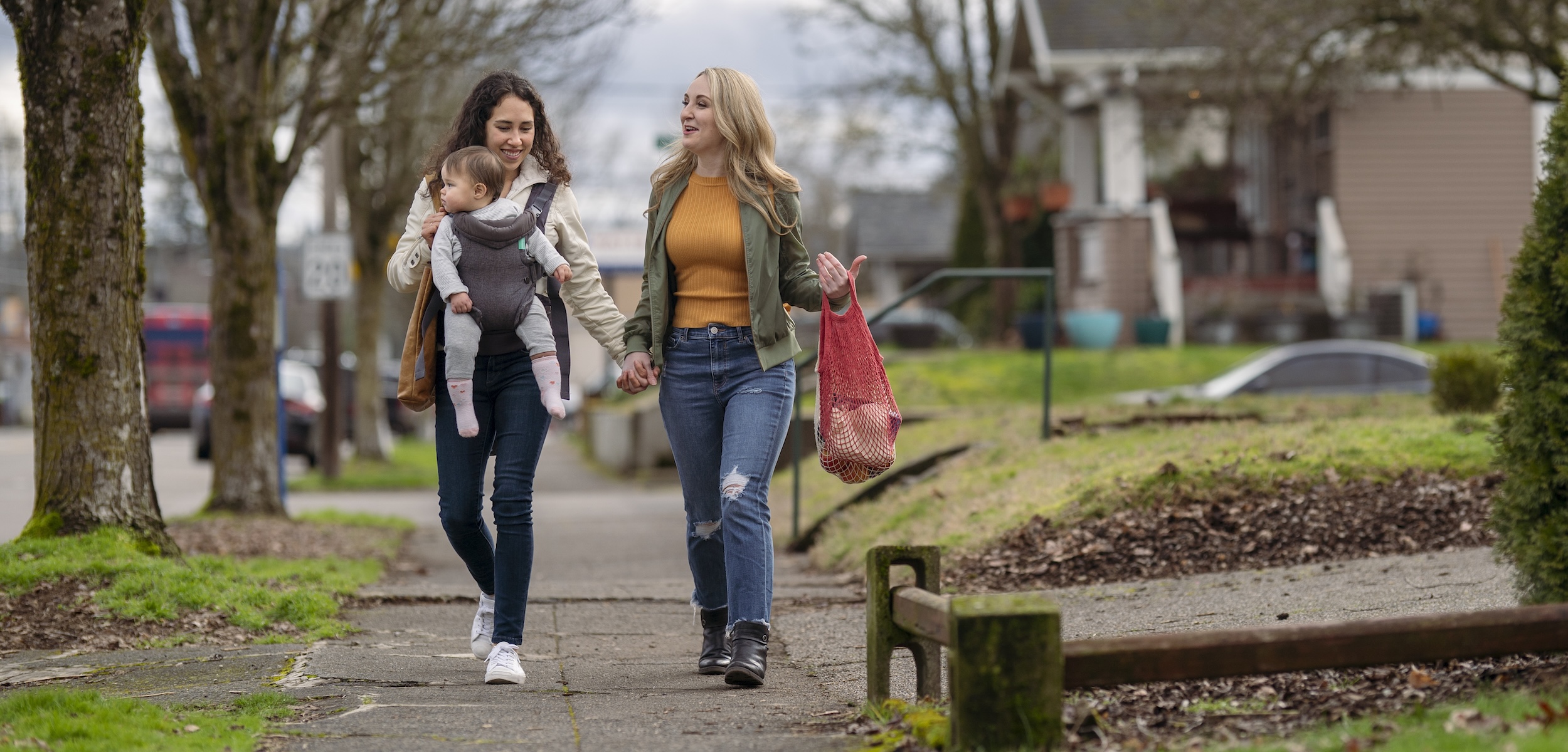 A young mixed race lesbian couple hold hands while walking on a sidewalk through an urban residential neighborhood. One woman is carrying a one year old baby in an ergonomic baby carrier while the other woman carries groceries in sustainable reusable shopping bags.