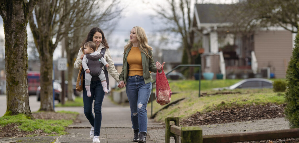 Same sex female couple holding hands while on a walk with baby girl A young mixed race lesbian couple hold hands while walking on a sidewalk through an urban residential neighborhood. One woman is carrying a one year old baby in an ergonomic baby carrier while the other woman carries groceries in sustainable reusable shopping bags.
