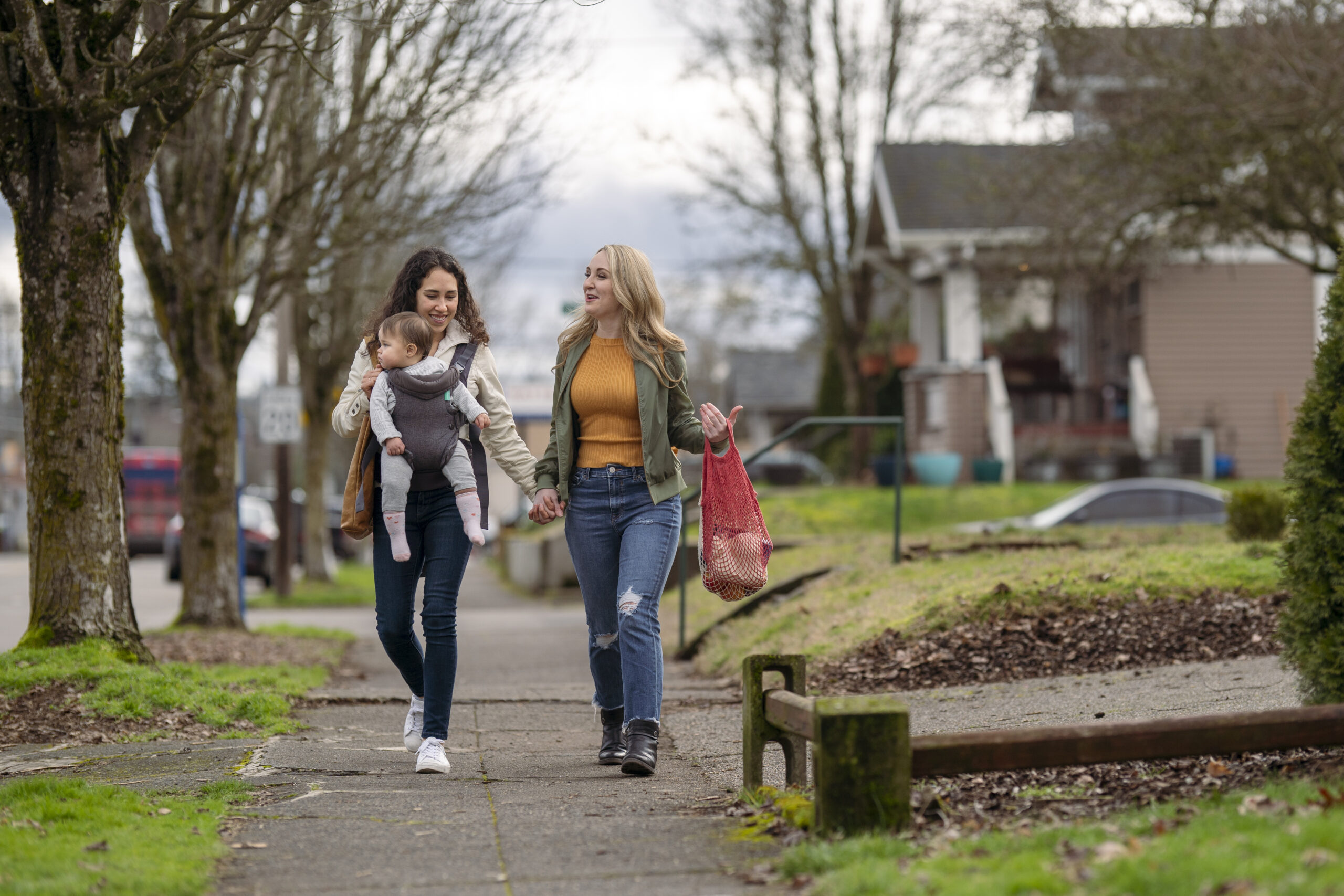 A young mixed race lesbian couple hold hands while walking on a sidewalk through an urban residential neighborhood. One woman is carrying a one year old baby in an ergonomic baby carrier while the other woman carries groceries in sustainable reusable shopping bags.
