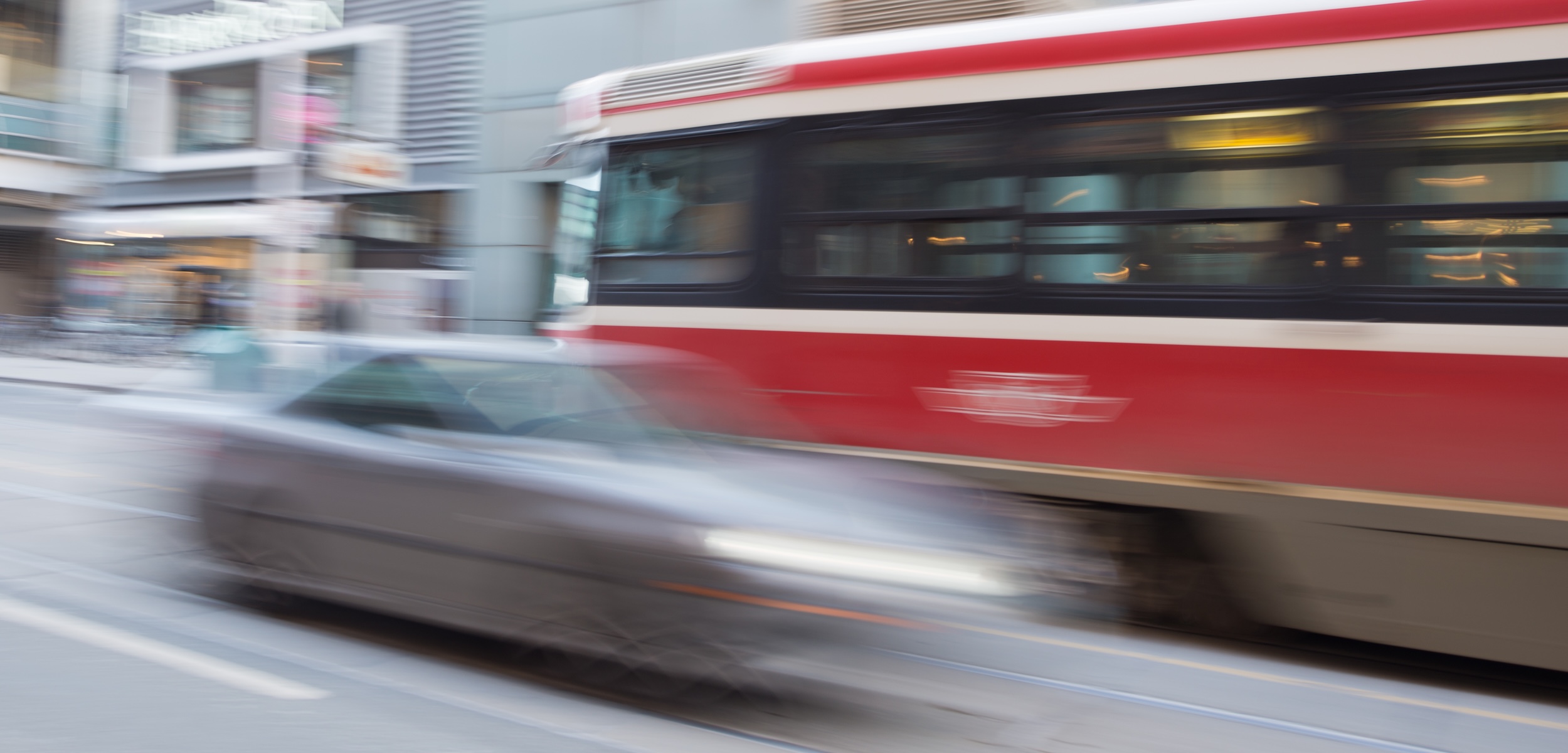 A blurry grey car speeding past a public bus in a downtown city