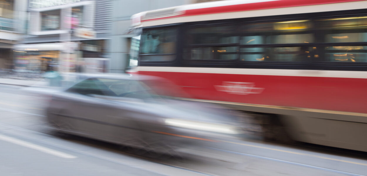 A blurry grey car speeding past a public bus in a downtown city