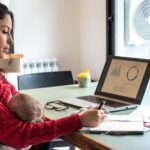 Young mom working home office on laptop while hold her baby on lap at home