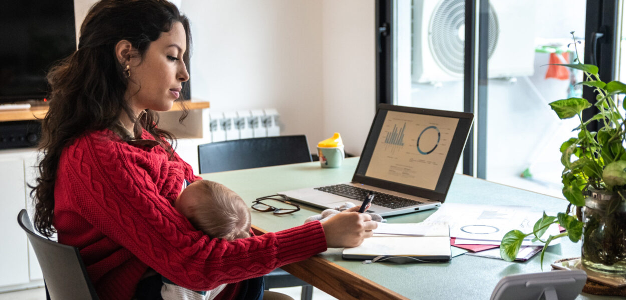 Young mom working home office on laptop while hold her baby on lap at home