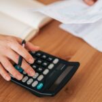 Person's hands using a calculator, holding bills, and taking notes on a wooden table, representing personal finance management