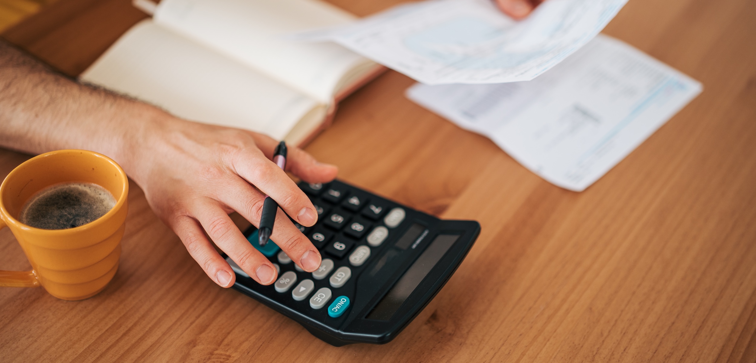 Person's hands using a calculator, holding bills, and taking notes on a wooden table, representing personal finance management